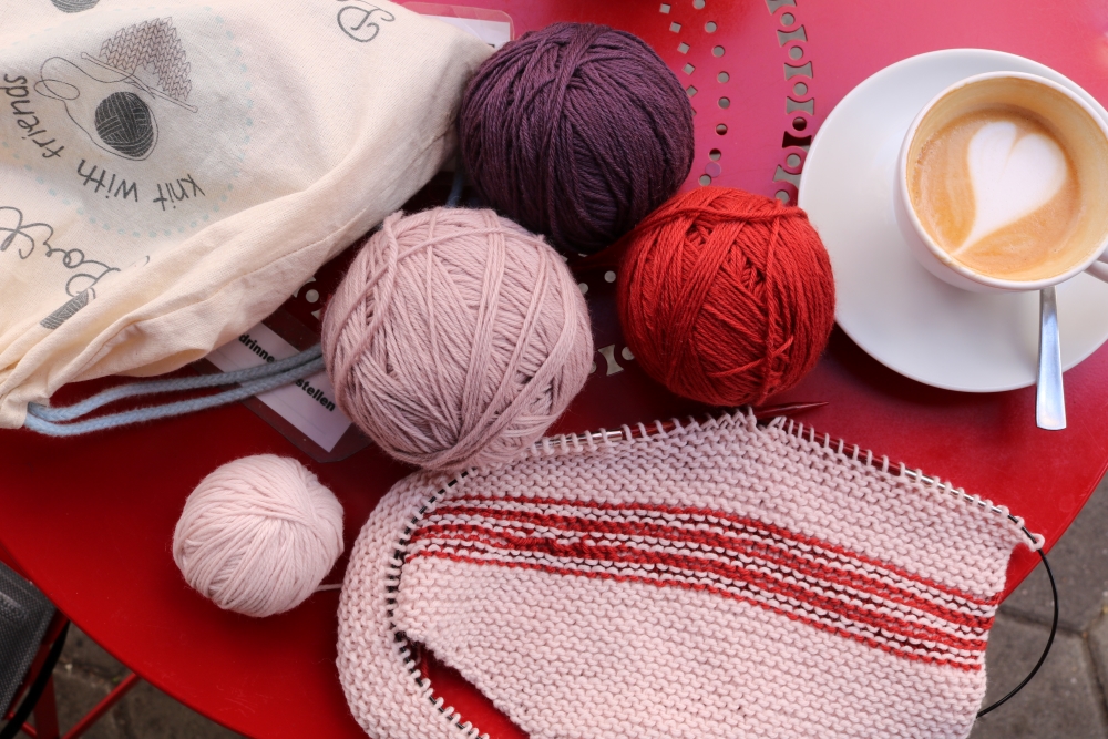 Different-coloured balls of yarn and an unfinished knitted piece lying on a table next to a cup of coffee